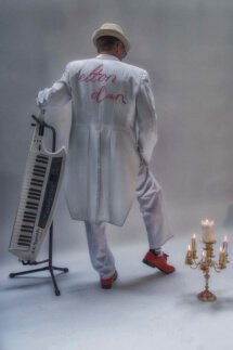 Elton Dan wearing red shoes, piano socks and a white suit with tails and "Elton Dan" embroidered on the back. A candlabra is on the floor to his right and he's leaning on his portable keyboard.