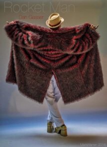 Elton Dan, wearing a tan hat, white pants, gold shoes and a red-tinged fur coat which he's holding open. His back is to the camera.