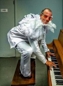Elton Dan backstage in white outfit with feathers, crouching on piano bench, playing piano.
