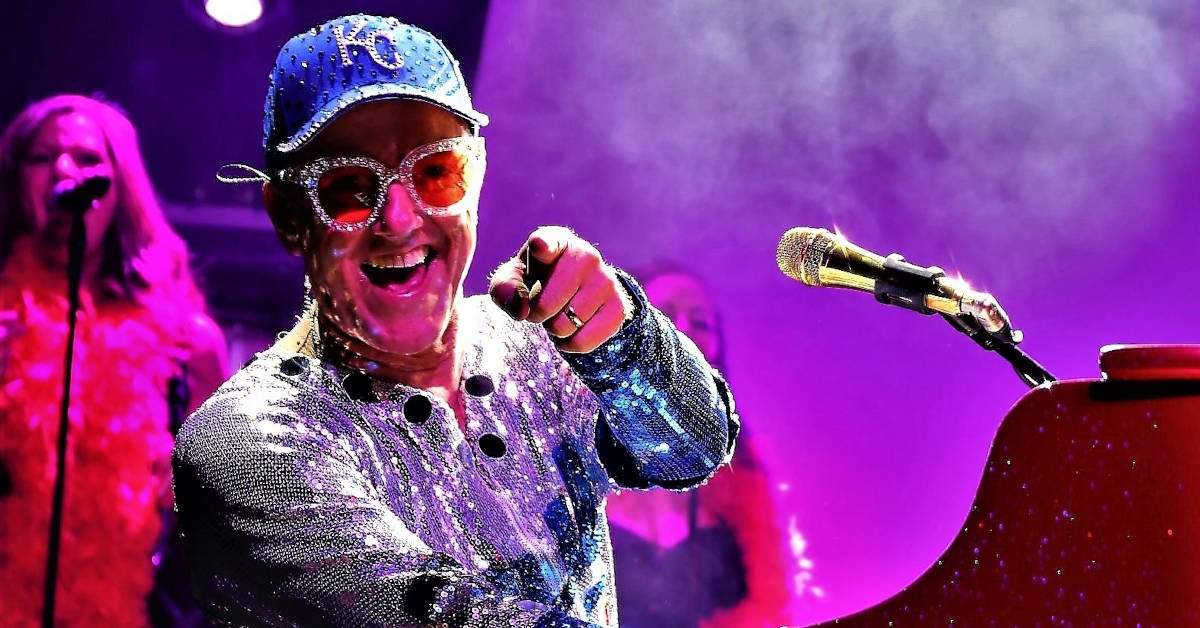 Elton Dan at his red piano, reaching for a high note with his right hand. He's wearing a black coat with tails that says, "Rocket Man" on the back.