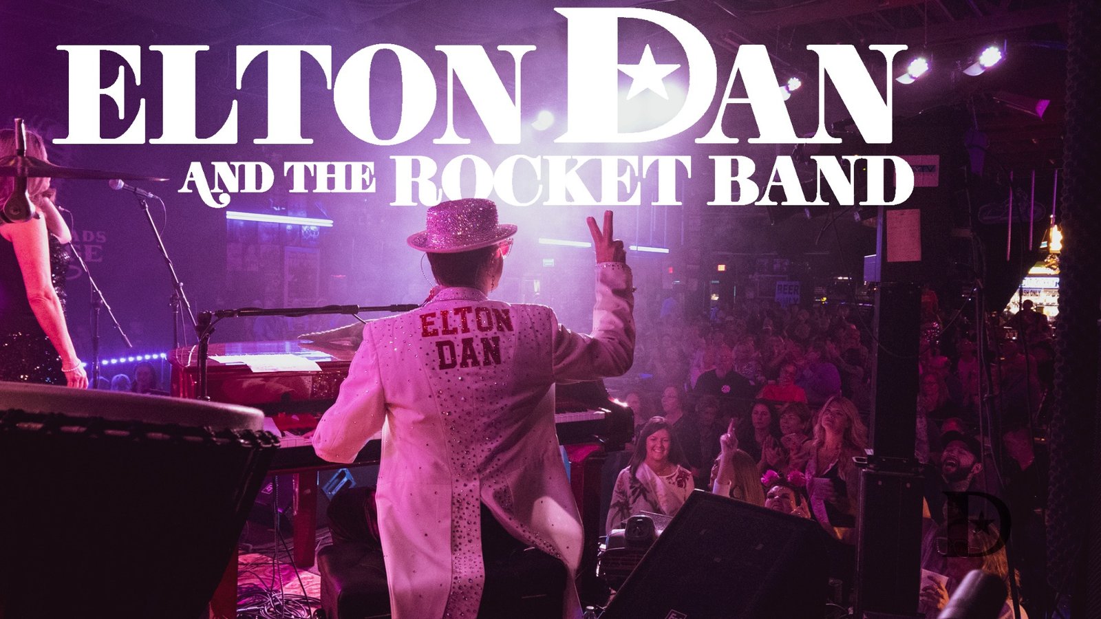 A shot of Elton Dan and the Rocket band on stage with the crowd in the foreground. White lights are shining beams of light in all directions from the stage.