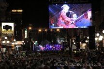 A shot from far back in the crowd at an Elton Dan street concert. A huge screen on a pole in the middle distance shows Dan in action in real time.