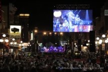 Photo from behind the crowd at a street concert, a large screen showing the action on stage is in the middle-ground.
