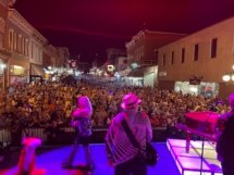 A shot from the stage of a large crowd at a street concert. The guitarist is in the center foreground, back turned and the backing vocalist is walking left.