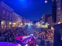 A beautiful shot of Dan playing his red piano on stage at a street concert with two spotlights in the distance and a large crowd in the background.