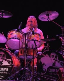 Elton Dan's drummer sitting behind his kit, which has black and white images of Elton Dan on the fronts of his two bass drums.