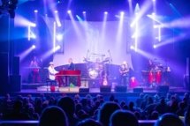 A shot of Elton Dan and the Rocket band on stage with the crowd in the foreground. White lights are shining beams of light in all directions from the stage.