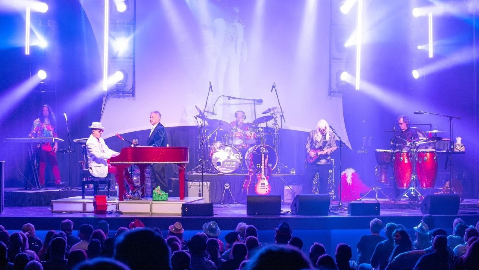 A shot of Elton Dan and the Rocket band on stage with the crowd in the foreground. White lights are shining beams of light in all directions from the stage.