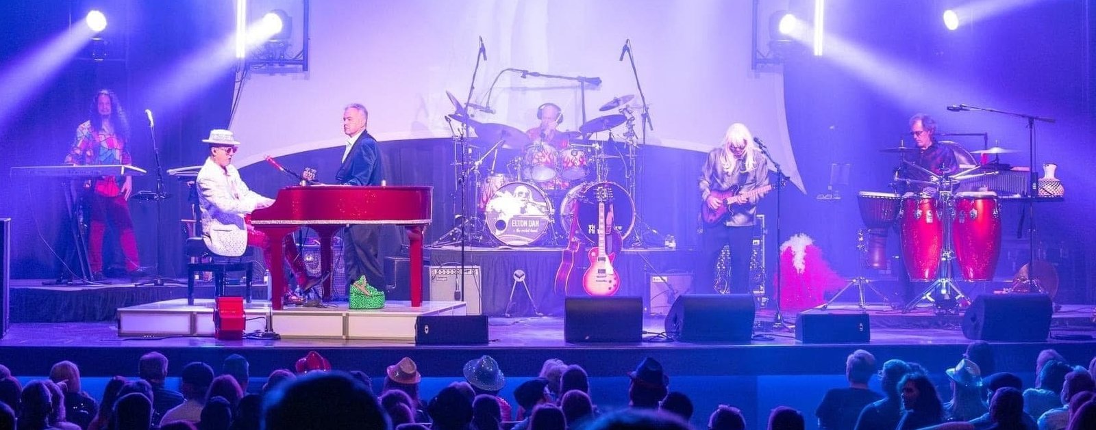 A shot of Elton Dan and the Rocket band on stage with the crowd in the foreground. White lights are shining beams of light in all directions from the stage.