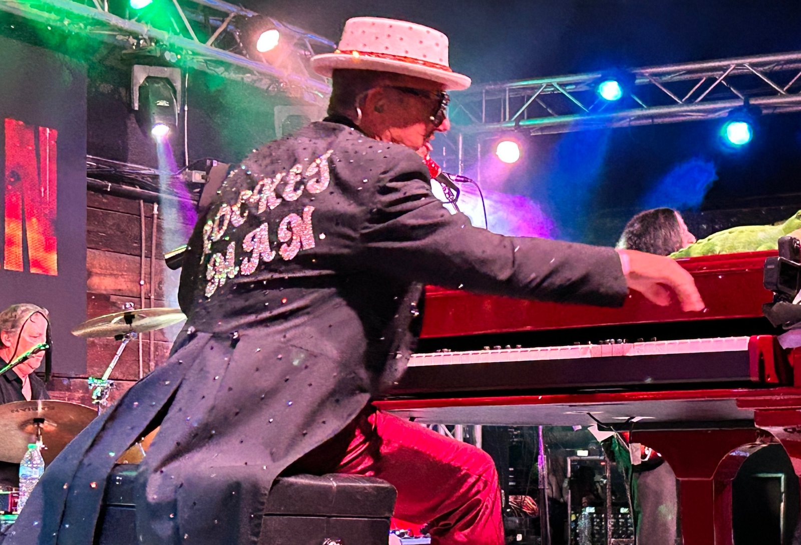Elton Dan at his red piano, reaching for a high note with his right hand. He's wearing a black coat with tails that says, "Rocket Man" on the back.