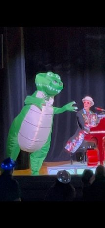 Elton Dan at his red piano with a man in a crocodile suit dancing next to him.