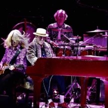 Dan playing piano on stage in a leopard print fur coat with guitarist rocking out on the bench with him. Percussionist is in the background.