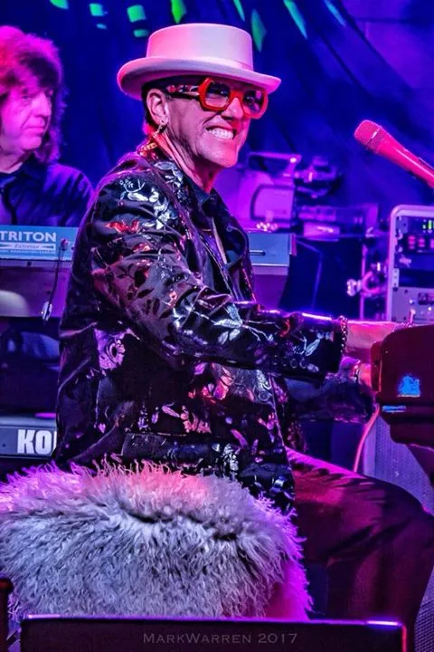 Elton Dan wearing a mirror jacket, hat and Elton glasses, sitting on a furry piano bench and grinning at the audience while he plays piano.