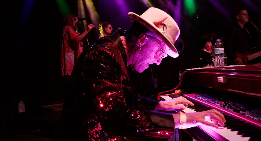 A shot of Elton Dan and the Rocket band on stage with the crowd in the foreground. White lights are shining beams of light in all directions from the stage.