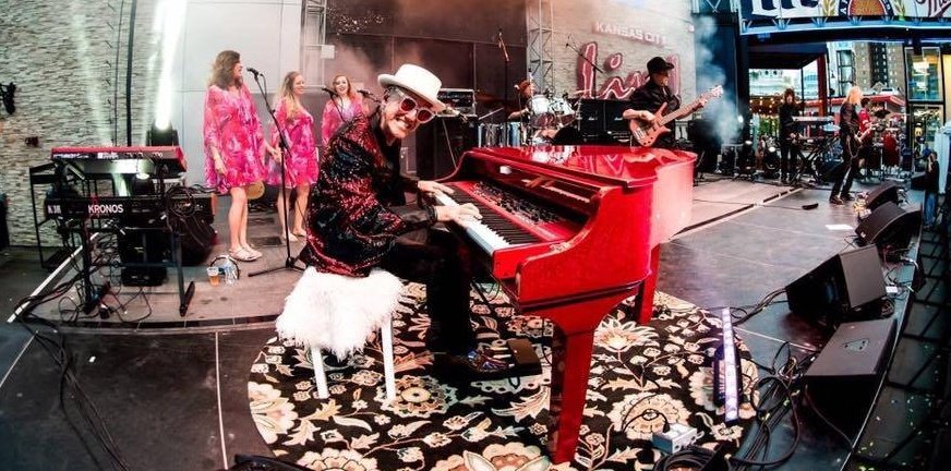 A shot of Elton Dan and the Rocket band on stage with the crowd in the foreground. White lights are shining beams of light in all directions from the stage.