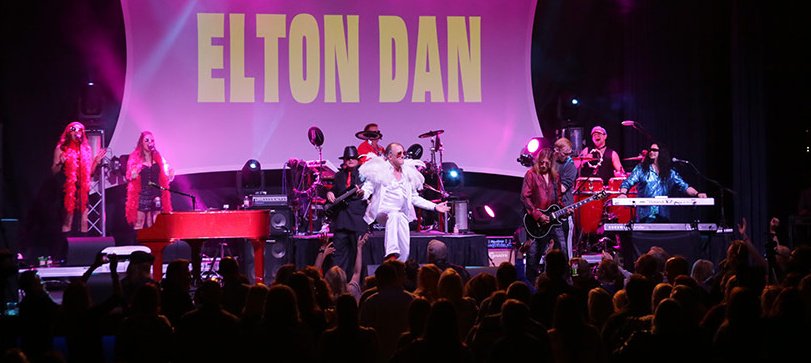 A shot of Elton Dan and the Rocket band on stage with the crowd in the foreground. White lights are shining beams of light in all directions from the stage.